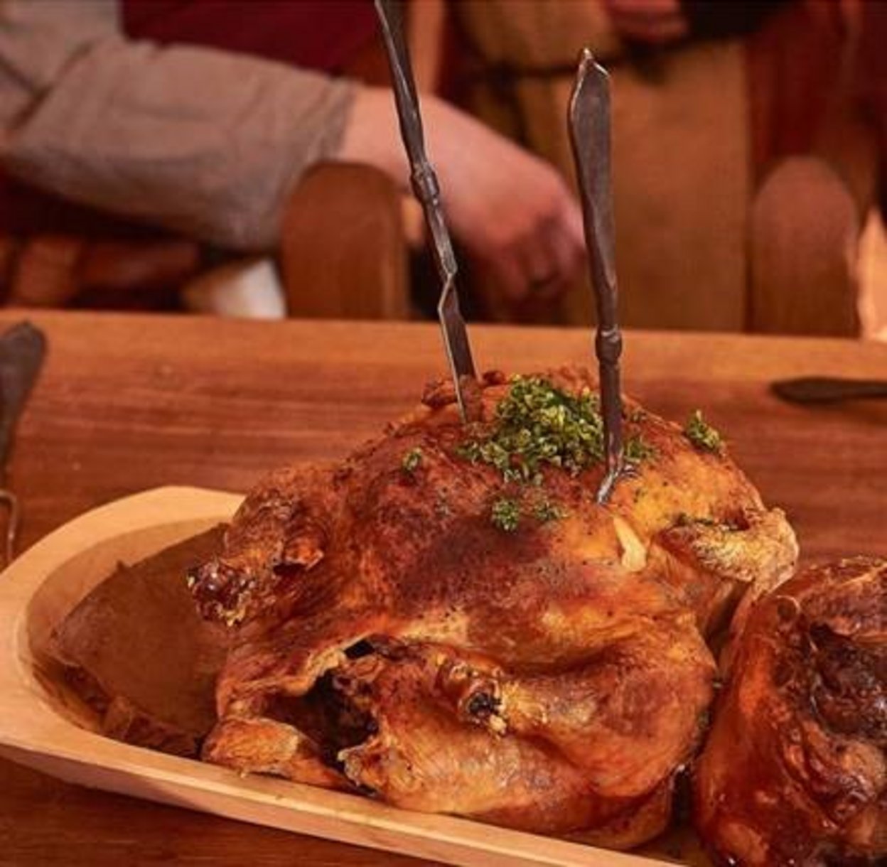 In einem zubereiteten Hühnchen in einer Holzschale stecken alte Messer und Gabel, steht auf einem Tisch, im Hintergrund sieht man den Arm eine rvemutlich sitzenden Person verschwommen