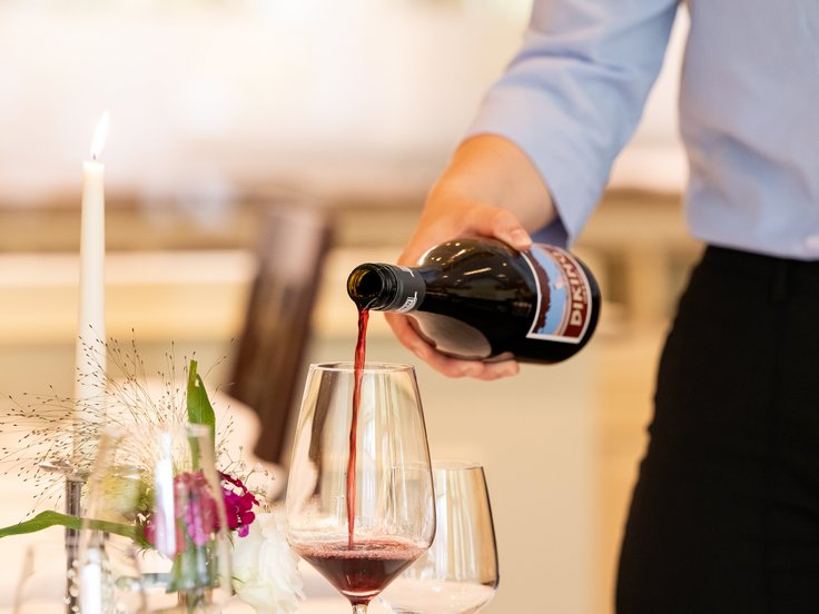 Gedeckter Tisch im Restaurant, Tiefer Telller mit Blumendeko, Person steht an der Seite und gießt vermutlich Rotwein aus einer Flasche in ein Glas