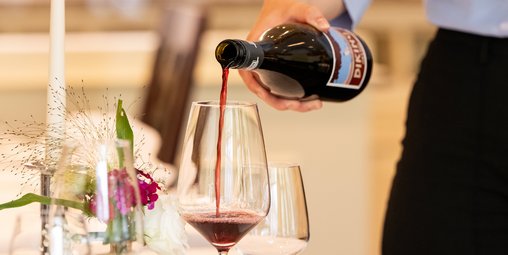 Gedeckter Tisch im Restaurant, Tiefer Telller mit Blumendeko, Person steht an der Seite und gießt vermutlich Rotwein aus einer Flasche in ein Glas