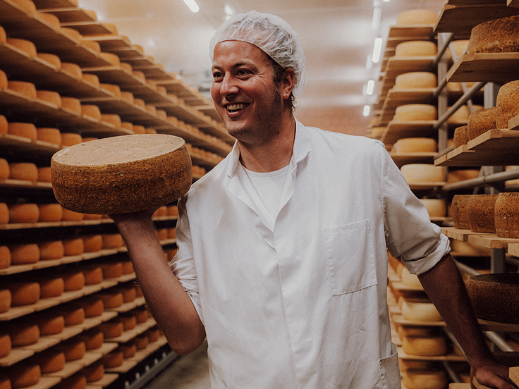 Käser Thilo hält einen Laib Käse im Käsekeller zwischen Regalen gefüllt mit weiteren Käselaiben in die Höhe und grinst