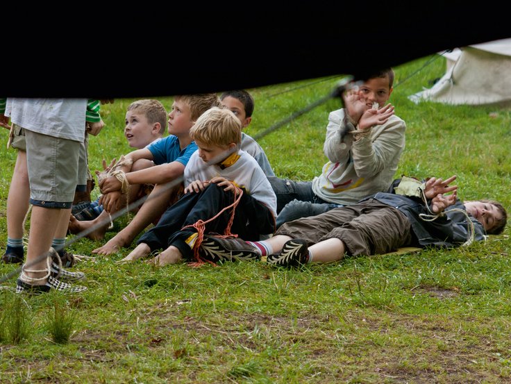 Gruppe von Kindern sitzt im Gras, viele haben Seile um Hände oder Füße gebunbden, zwei zeigen die Hände in die Kamera, Rest schaut auf zu zwei weiteren Kindern, die vor der sitzenden Kindergruppe stehen