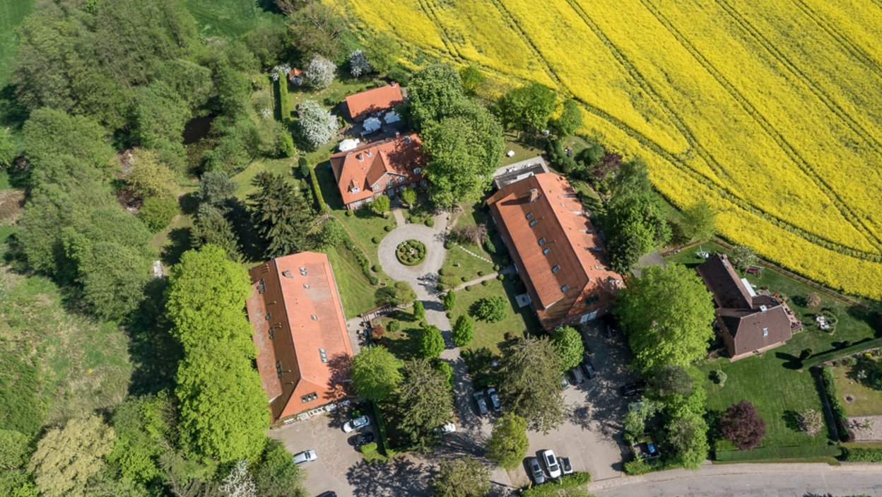 Luftbildaufnahme, in der Mitte ein drei seiten Hof, rechts ein weiteres Gebäude, davor PArkplätze, ringsum viel Grün aus Bäumen und Feldern, auf einem Feld blüht vermutlich Raps leuchtend gelb