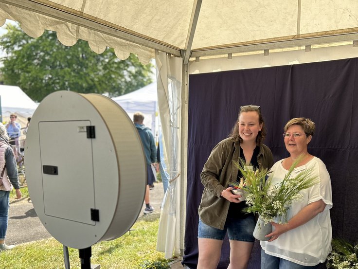 Zwie Personen lächeln für ein Foto an der Fotobox in die Kamera. In der Hand halten sie Pflanzen und Obst sowie Gemüse. Der Hintergrund der Fotobox ist blau. Im Hintergrund ist Veranstaltungstreiben. 