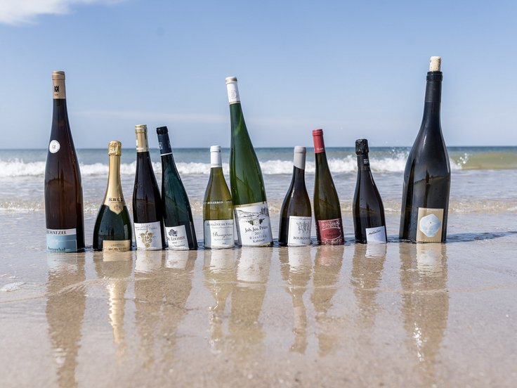 Mehrere verschiedene Flaschen Wein stehen am Strand, dahinter das Meer mit Wellen