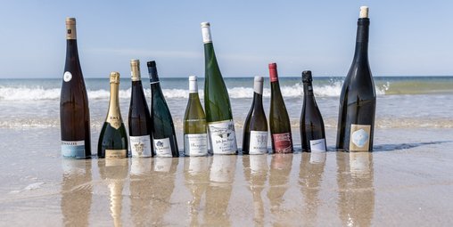 Mehrere verschiedene Flaschen Wein stehen am Strand, dahinter das Meer mit Wellen