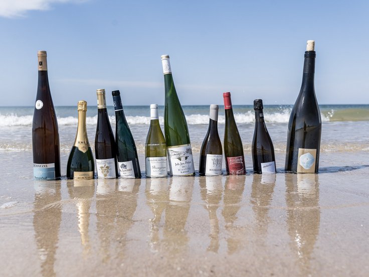 Mehrere Weinflaschen stehen im Sand vor dem Meer