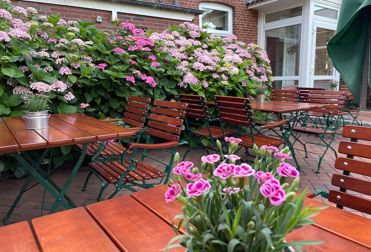 Außenbereich gepflastert mit Tischen und Stühlen, Dekopflanzen auf Tischen, dahinte rgroße Hortensienbüsche, rechts der Eingangsbereich aus Glas mit weißen Rahmen und ein Sonnenschirm zusammengeklappt