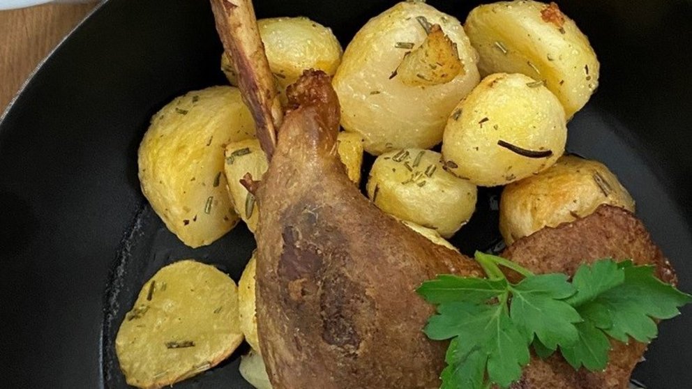 In Pfanne ist Gericht mit Bratkartoffeln und Gänsekeule angerichtet, daneben steht ein Gefäß mit dunkler Soße